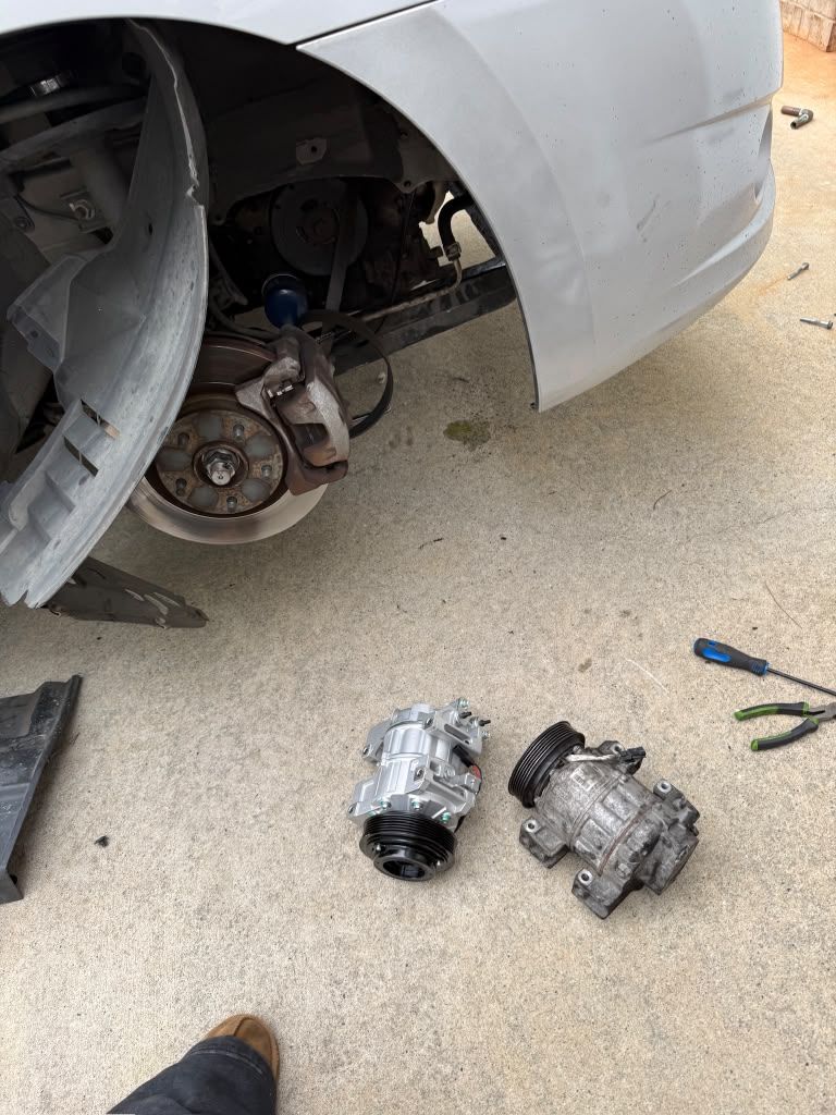 A new and an old A/C compressor on the ground next to a car with the front wheel removed