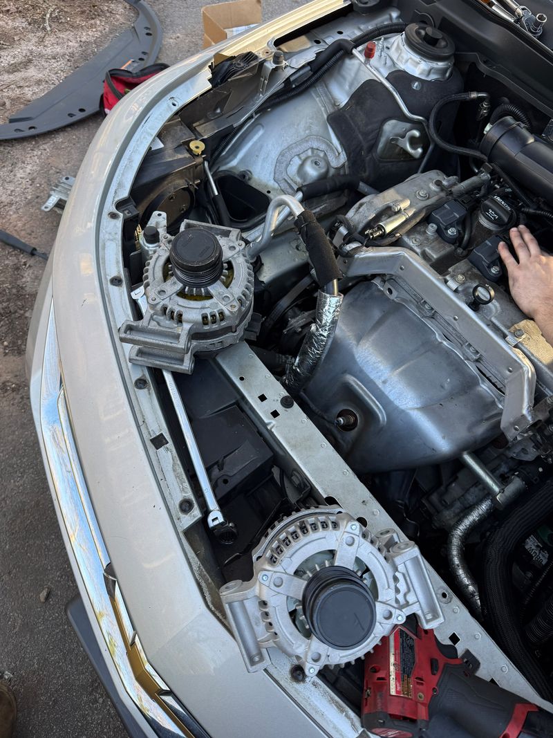 An old and a new alternator resting on the front of a car with the engine bay open