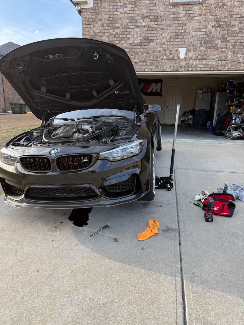 BMW M4 with the hood open on a floor jack, drained oil on the driveway, tools beside it