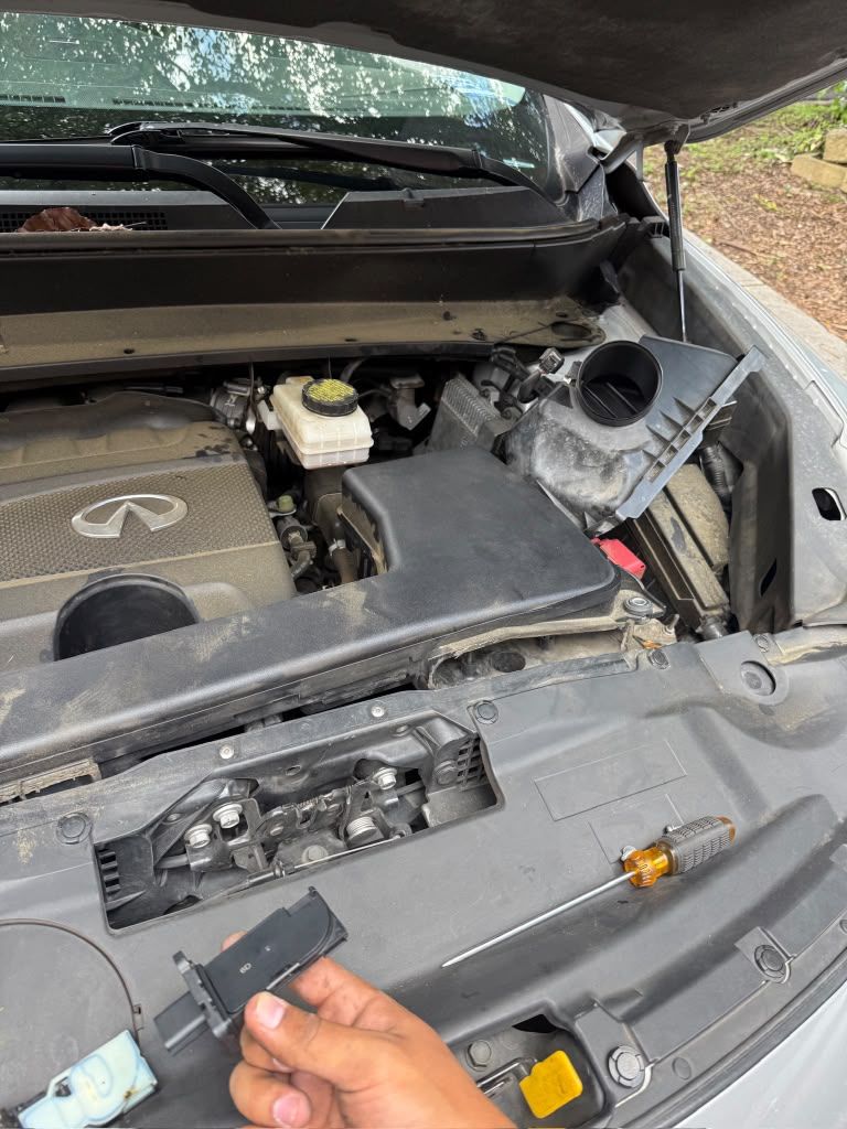 Infiniti engine bay with the air intake opened and a technician holding the removed sensor